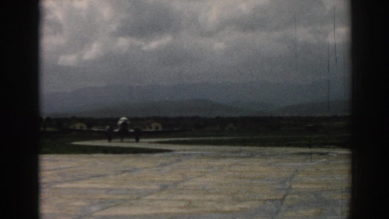 Small silver aircraft taxis along the Izmir runway area, with low buildings or shapes and mountains beyond.
