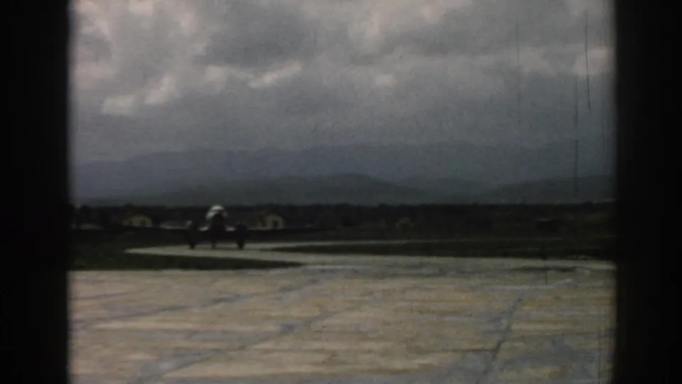 Small silver aircraft taxis along the Izmir runway area, with low buildings or shapes and mountains beyond.