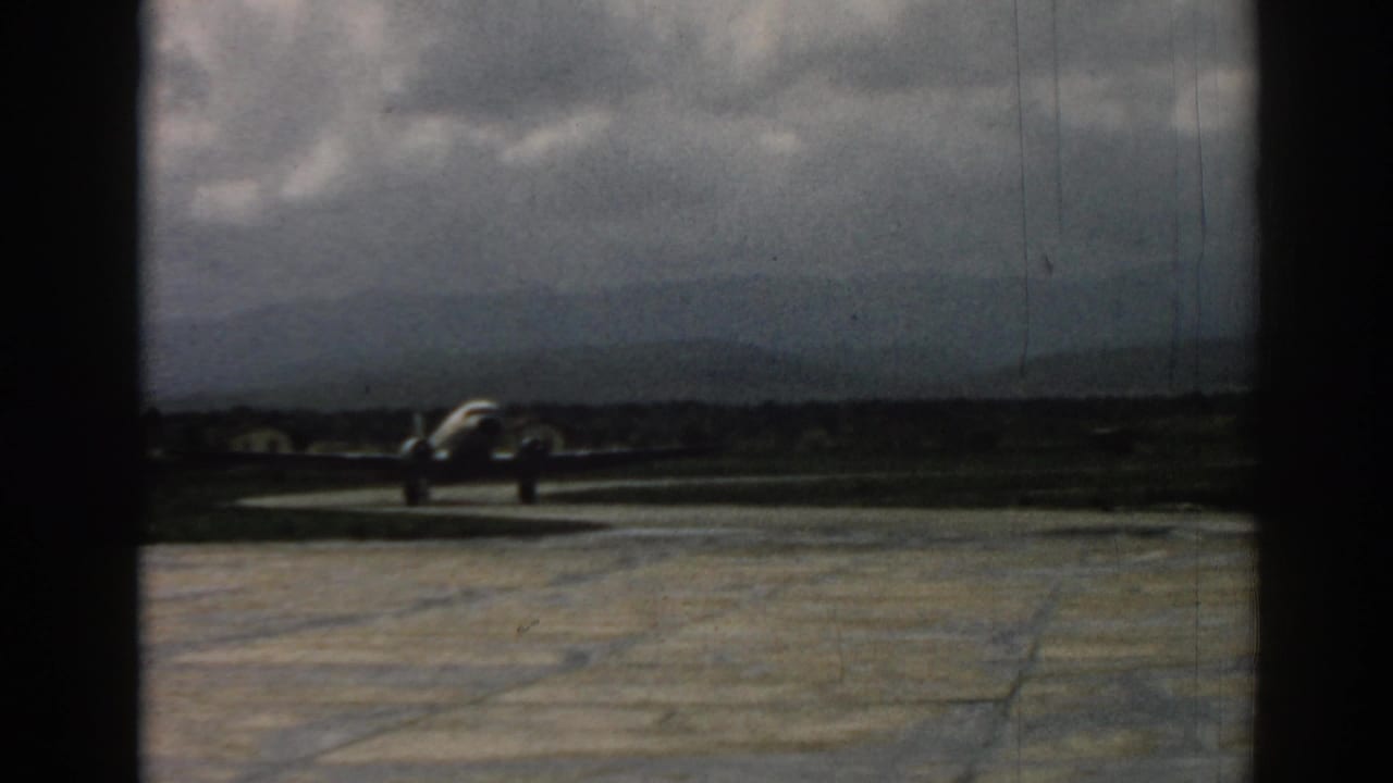 A silver aircraft taxis across the runway at the Izmir airfield, framed by dark film or window edges and distant hills.
