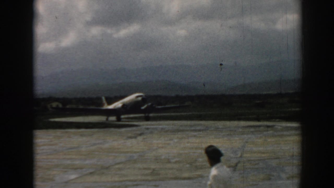 1962 Izmir airport scene with a propeller aircraft on the tarmac and a man walking through the foreground.