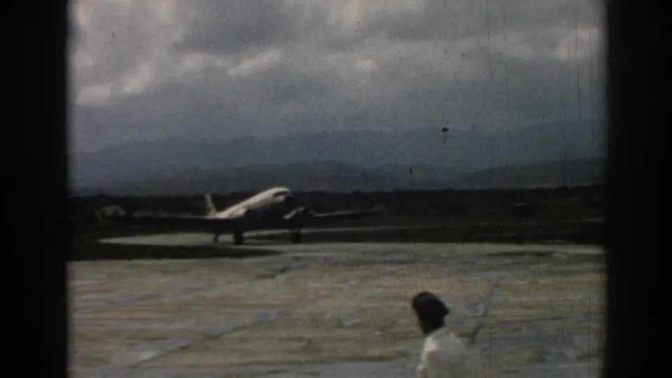 1962 Izmir airport scene with a propeller aircraft on the tarmac and a man walking through the foreground.