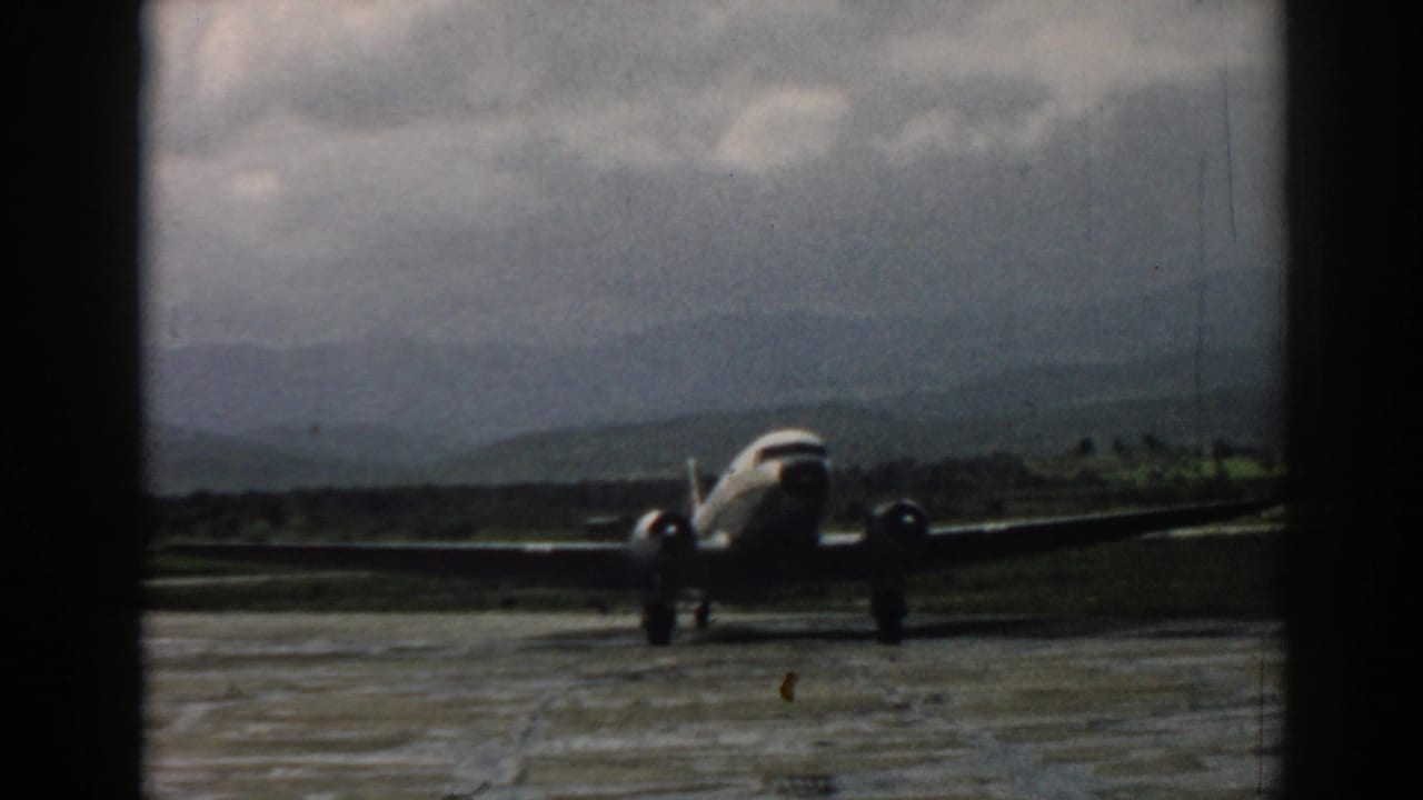 1962 Izmir home-movie frame showing a twin-engine propeller airplane moving on a wet aerodrome with hills in the distance.