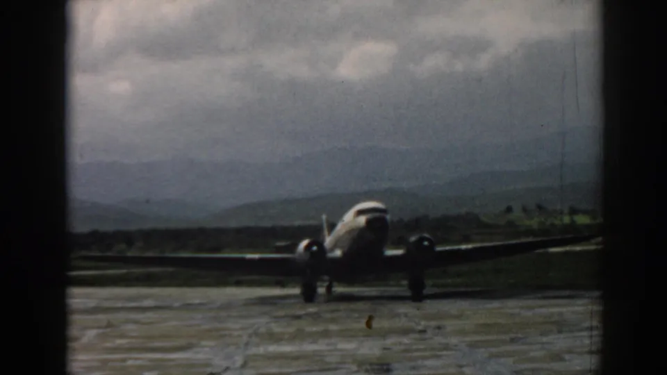 1962 Izmir home-movie frame showing a twin-engine propeller airplane moving on a wet aerodrome with hills in the distance.