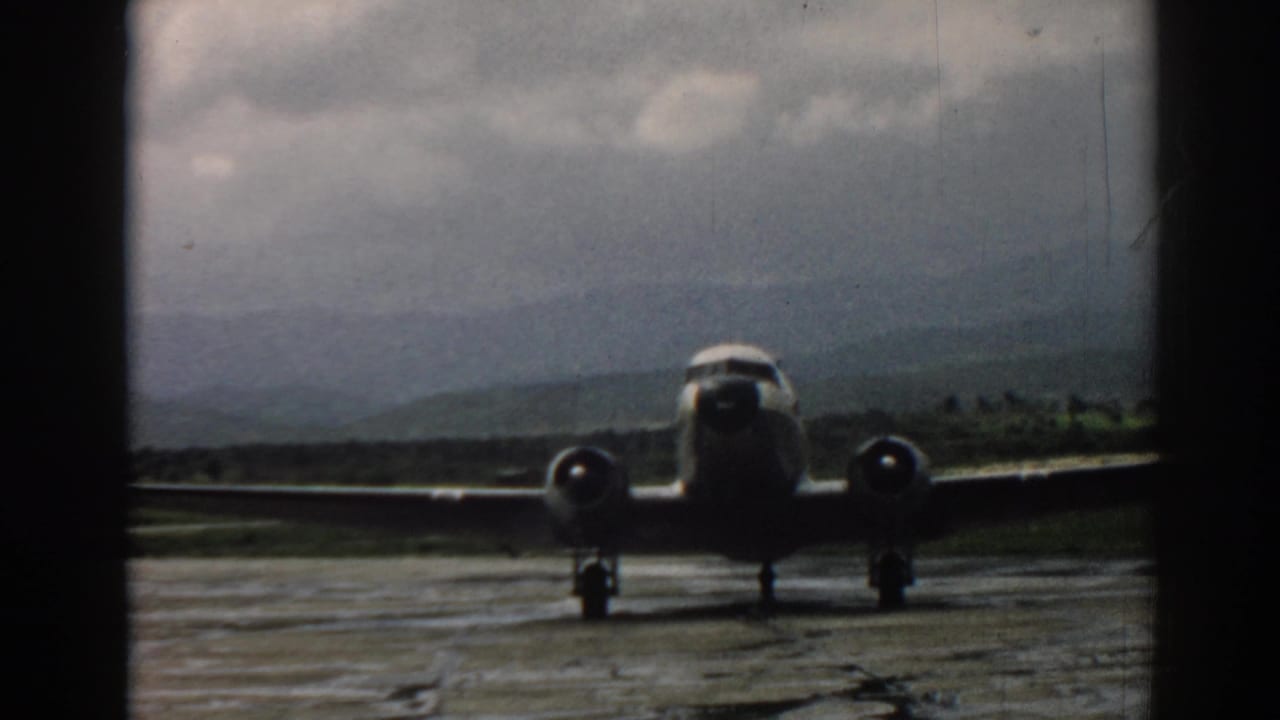 1962 Izmir runway view of a propeller airliner facing the camera through a dark window frame, with hills and clouded sky behind it.