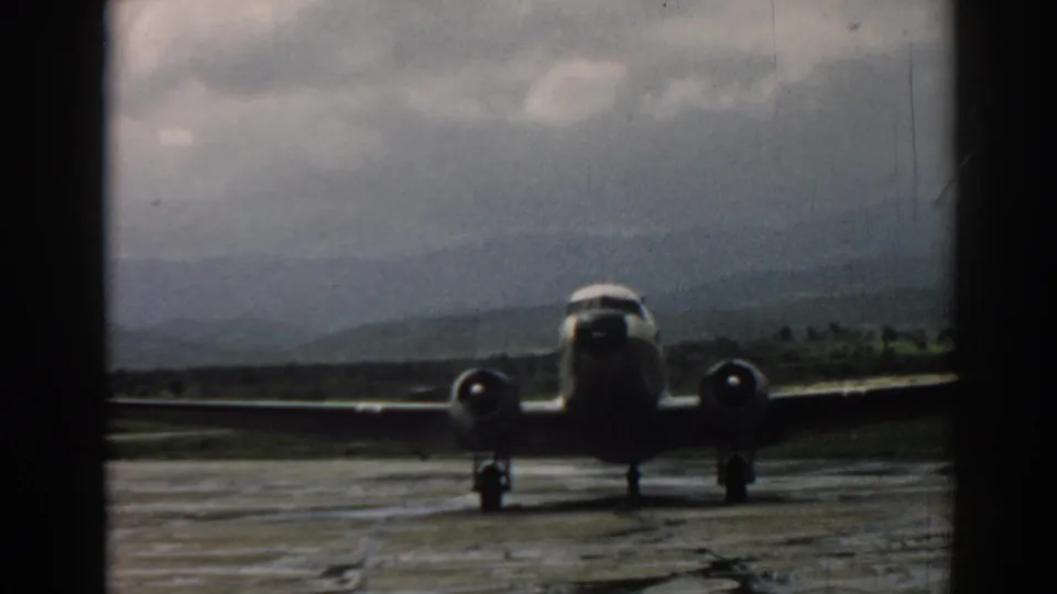 1962 Izmir runway view of a propeller airliner facing the camera through a dark window frame, with hills and clouded sky behind it.