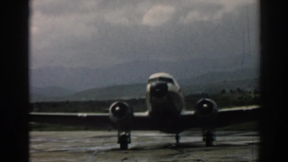 A 1962 Izmir airport view shows a propeller aircraft on the runway, filmed through a dark vertical window frame under a cloudy sky.