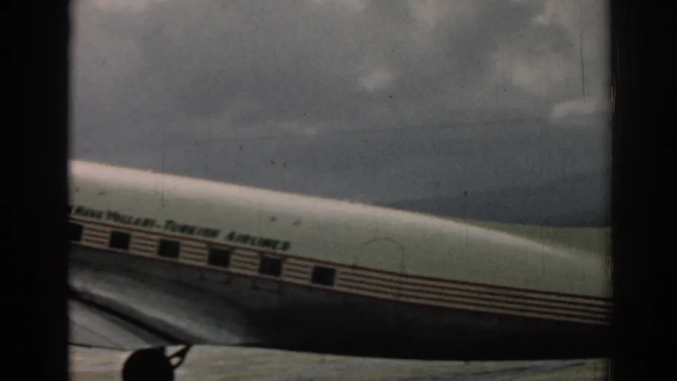 1962 Izmir airport footage shows the aircraft midsection moving past, with airline lettering, red stripes, wing, and landing gear in view.