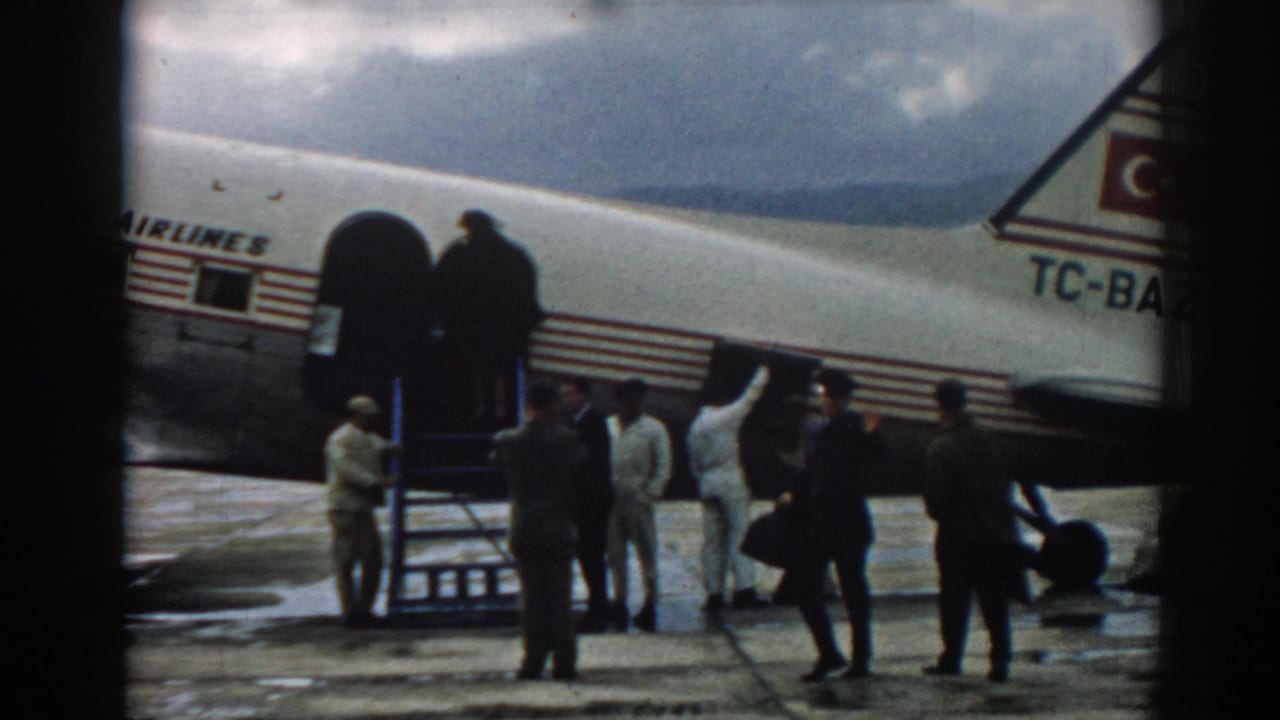 Uniformed and civilian men stand near Turkish Airlines boarding stairs on a wet Izmir tarmac in 1962.
