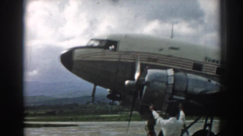 1962 Izmir home-movie frame showing a Turk Hava Yolları propeller aircraft nose, engine, cloudy sky, and ground crew member.