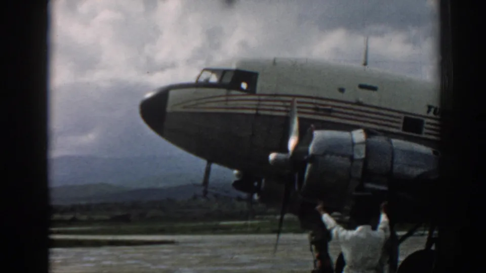 1962 Izmir frame of a Turkish Airlines propeller plane, showing the nose, engine nacelle, landing gear, cloudy sky, and directing crew member.