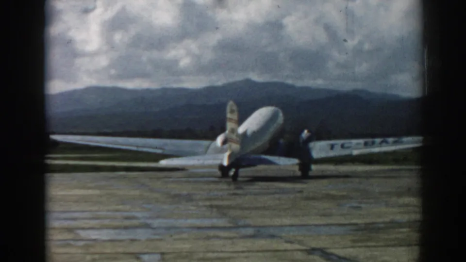 Wide 1962 Izmir airport view of a propeller aircraft on the tarmac, with a small ground figure, cloudy sky, and mountains behind.