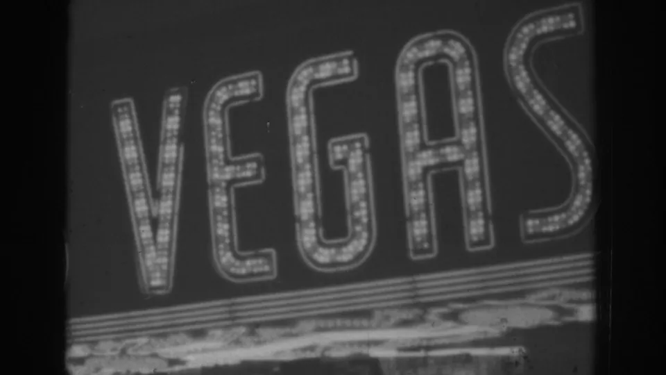 Black-and-white 1962 Las Vegas home-movie frame showing a close drive-by view of large illuminated VEGAS letters against the night.