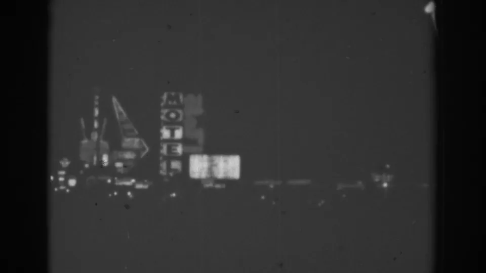 Black-and-white Las Vegas night frame showing a vertical motel sign, illuminated billboard panels, and distant roadside lights from a passing vehicle.