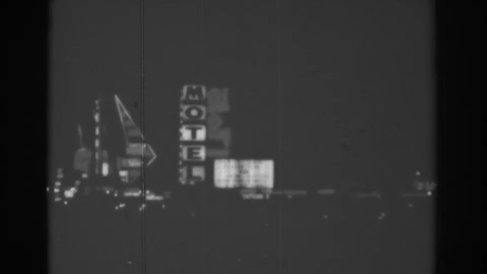 1962 Las Vegas night drive frame showing a vertical MOTEL sign, arrow-shaped neon, and blurred illuminated signs in black-and-white 8mm.