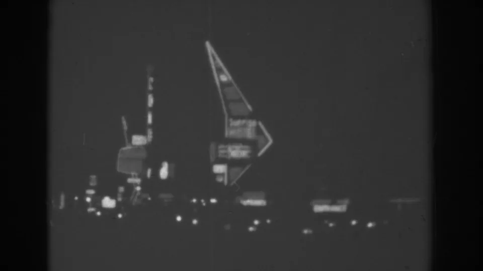 1962 Las Vegas home-movie frame showing a moving nighttime view of an arrow-shaped neon sign, vertical motel-area signage, and low roadside lights.
