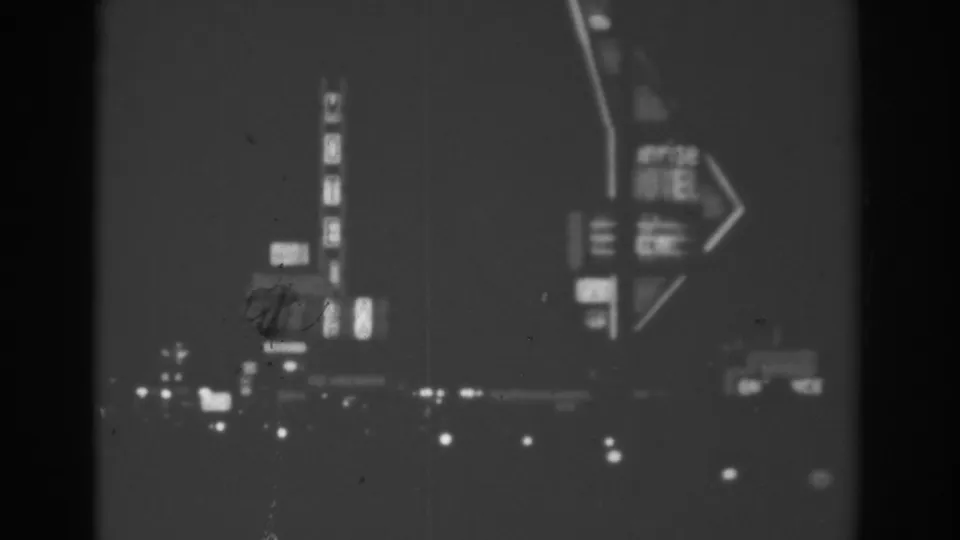 Black-and-white 8mm frame from a 1962 Las Vegas night drive, with a large arrow-shaped neon sign and glowing roadside lights.