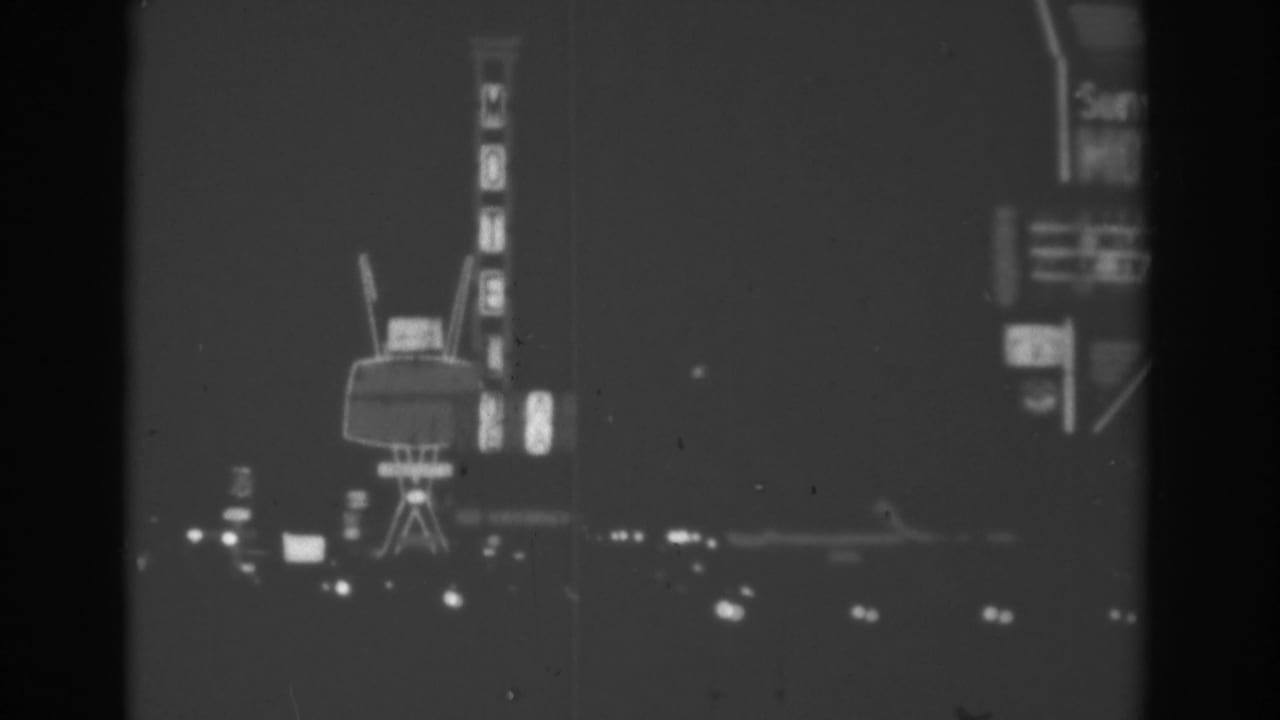 1962 Las Vegas night drive frame showing vertical motel signage, a large roadside sign, and blurred lights in black-and-white 8mm.