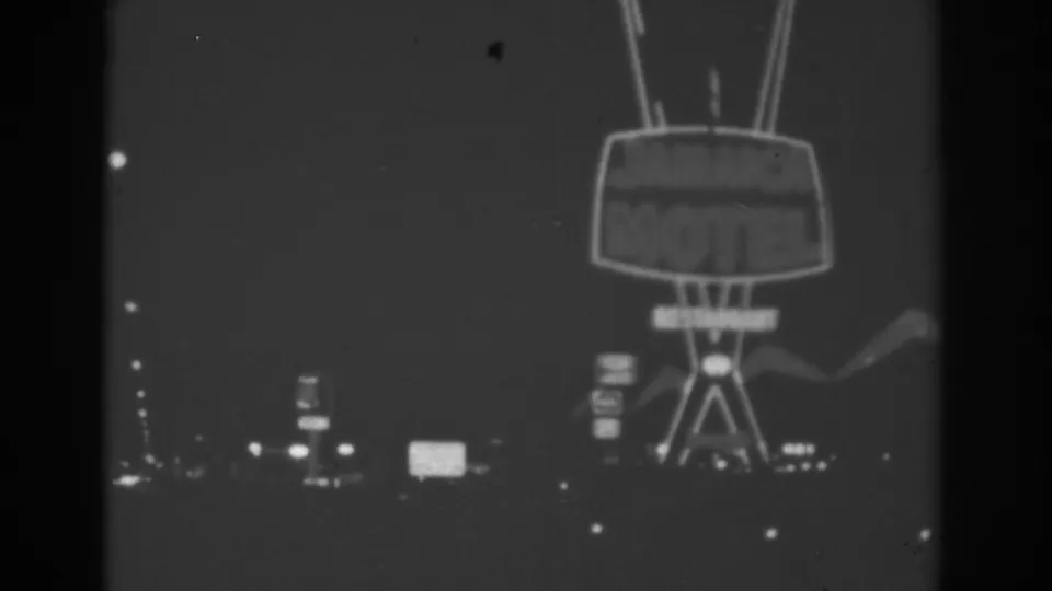 Black-and-white 1962 Las Vegas frame centered on a large retro motel sign, with distant signs and roadway lights below.