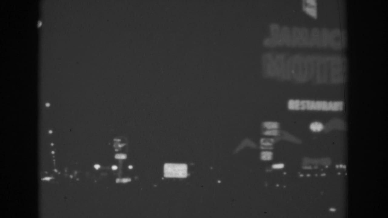 1962 Las Vegas night drive frame with soft black-and-white motel and roadside signage passing in the distance.