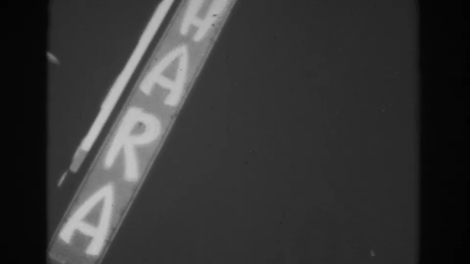 Close handheld view of a bright diagonal sign in 1962 Las Vegas, with large letters softened by black-and-white film exposure.