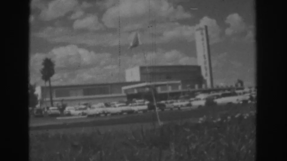 1962 Las Vegas daytime frame showing a low commercial building, flagpole, palm tree, and classic cars along the road.