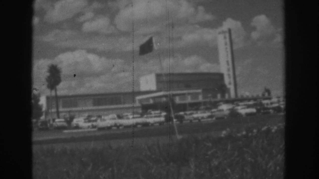 1962 Las Vegas home-movie view of the Dunes exterior with a waving flag, tall sign tower, palm tree, and cars along the frontage.