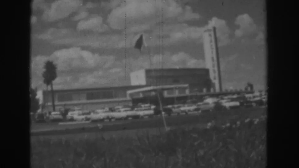 Black-and-white 8mm frame of the Dunes frontage in Las Vegas, showing a flag, sign tower, palm tree, and parked cars.