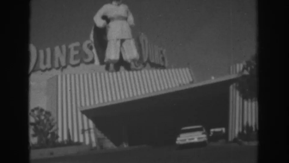 Black-and-white Las Vegas home-movie frame of the Dunes facade with rooftop figure, visible lettering, corrugated siding, and cars near the entrance.