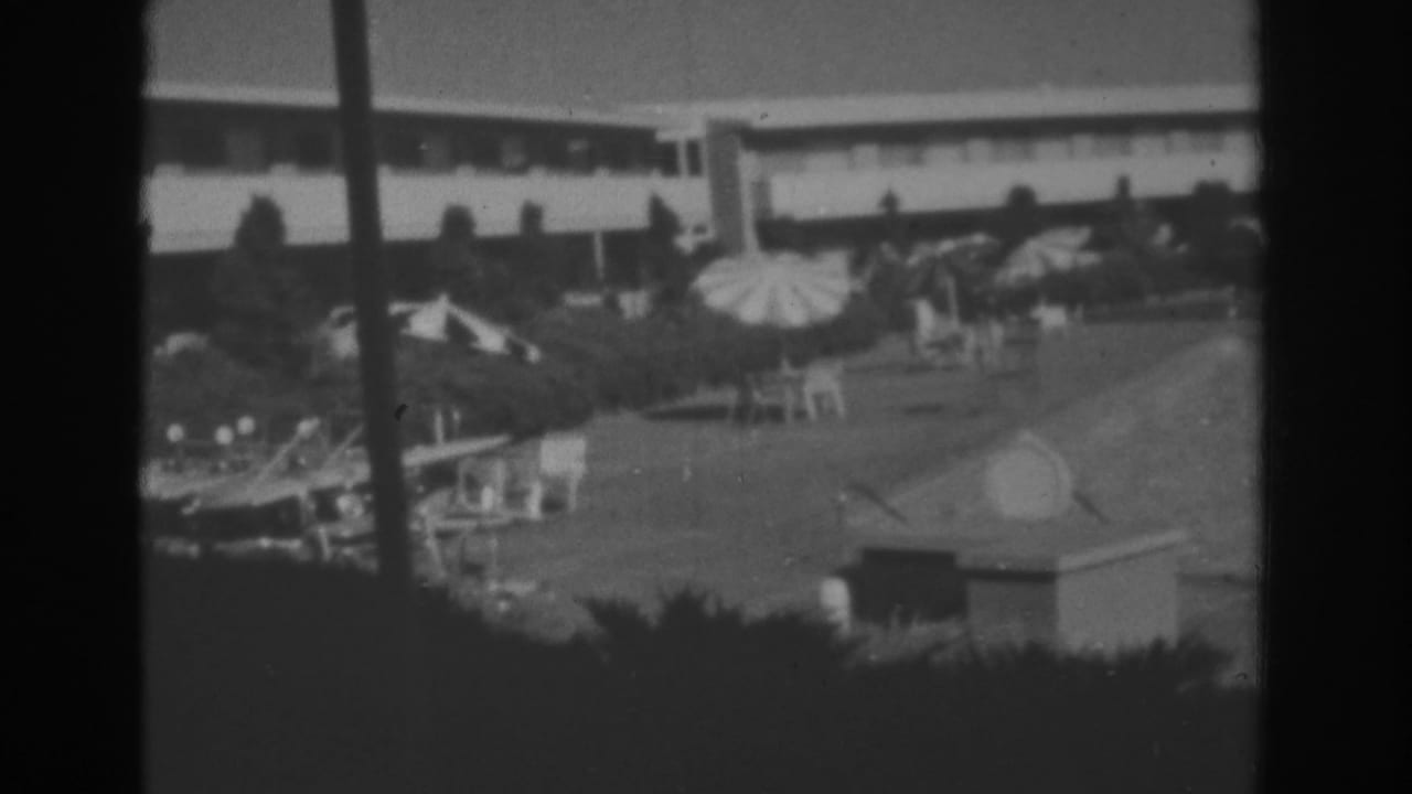 Distant motel courtyard with swimming pool, umbrellas, lounge chairs, hedges, and a low building.