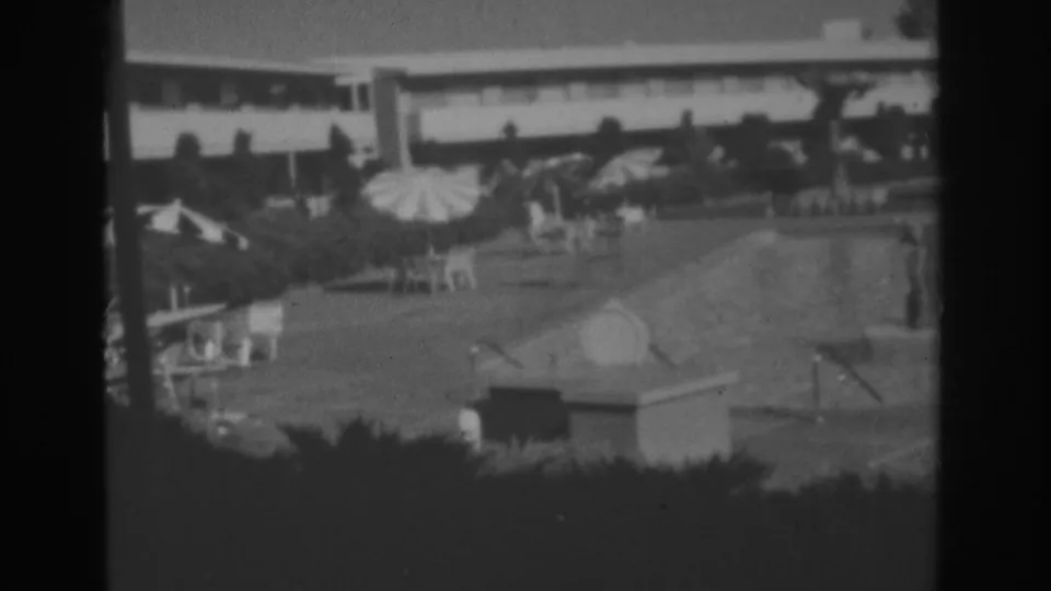 1962 Las Vegas home-movie frame of a motel pool area with patterned umbrellas, lounge chairs, hedges, and distant people near the pool.