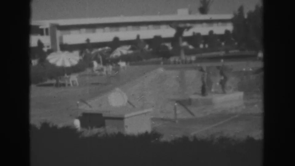 1962 Las Vegas 8mm frame showing a motel pool courtyard with guests in the water, an umbrella, lounge chairs, hedges, and a low building.