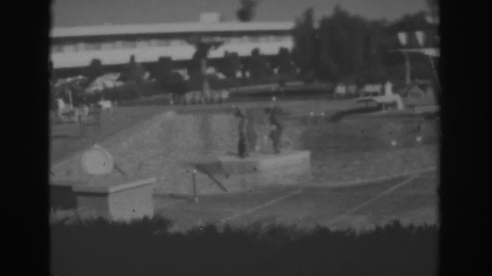 1962 Las Vegas home-movie frame of a motel or resort pool with distant guests in the water, umbrellas, chairs, hedges, trees, and a low building.