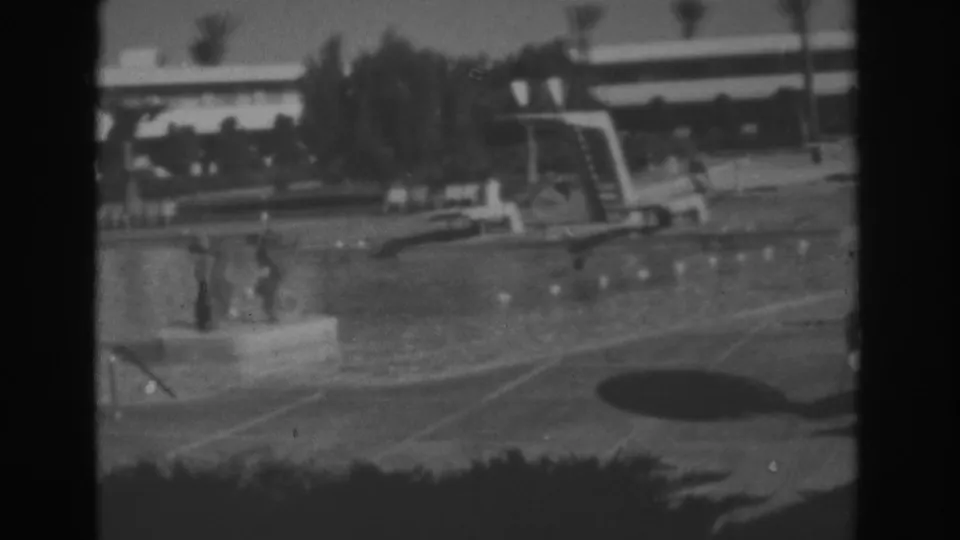 Black-and-white 8mm view of a Las Vegas hotel pool in 1962, with distant guests, a diving board, palm trees, and modern resort architecture.