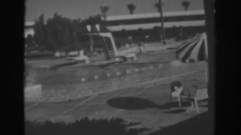 Las Vegas hotel pool area in 1962 with a striped umbrella, lounge chairs, diving board, palms, and soft black-and-white home-movie texture.