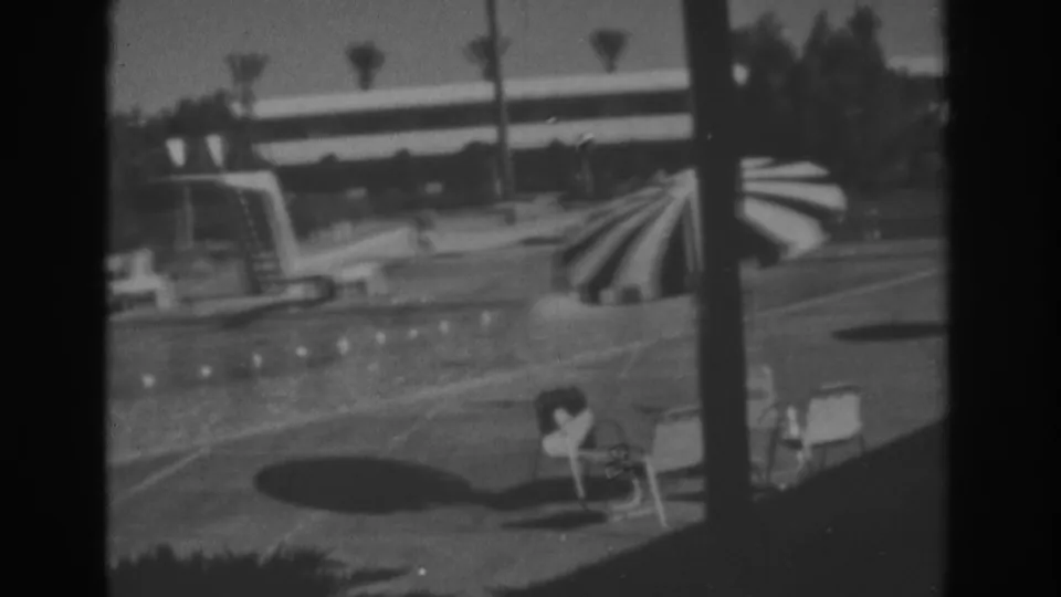 1962 Las Vegas resort poolside view showing a striped umbrella, lounge chairs, diving board, palm trees, and hotel structures in 8mm black-and-white footage.