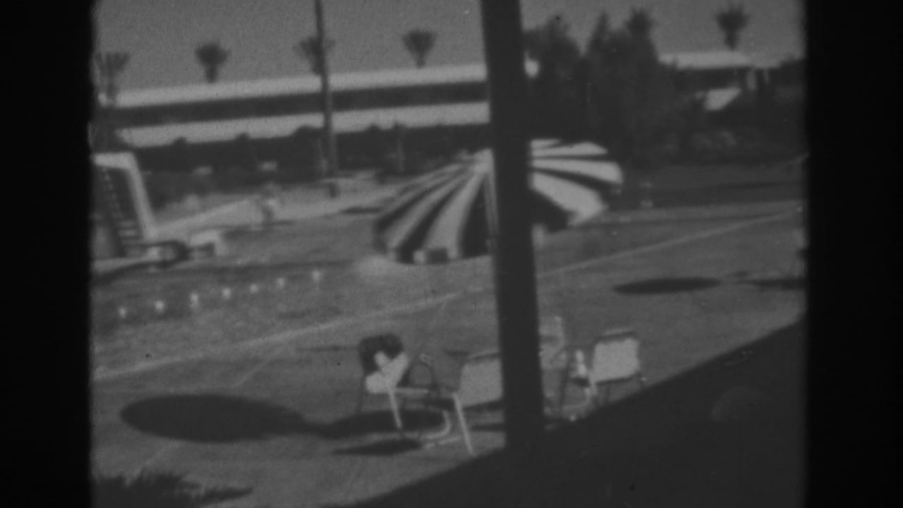 Resort swimming pool with striped umbrella, lounge chairs, palms, and a person near the deck.