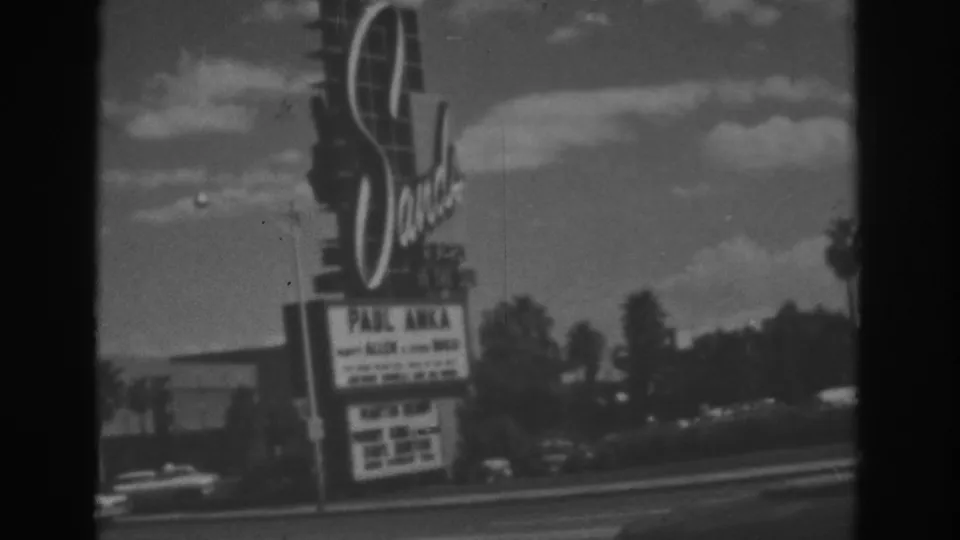 1962 Las Vegas home-movie frame with the Sands Hotel sign, Paul Anka marquee, cars, palm trees, and low roadside buildings.