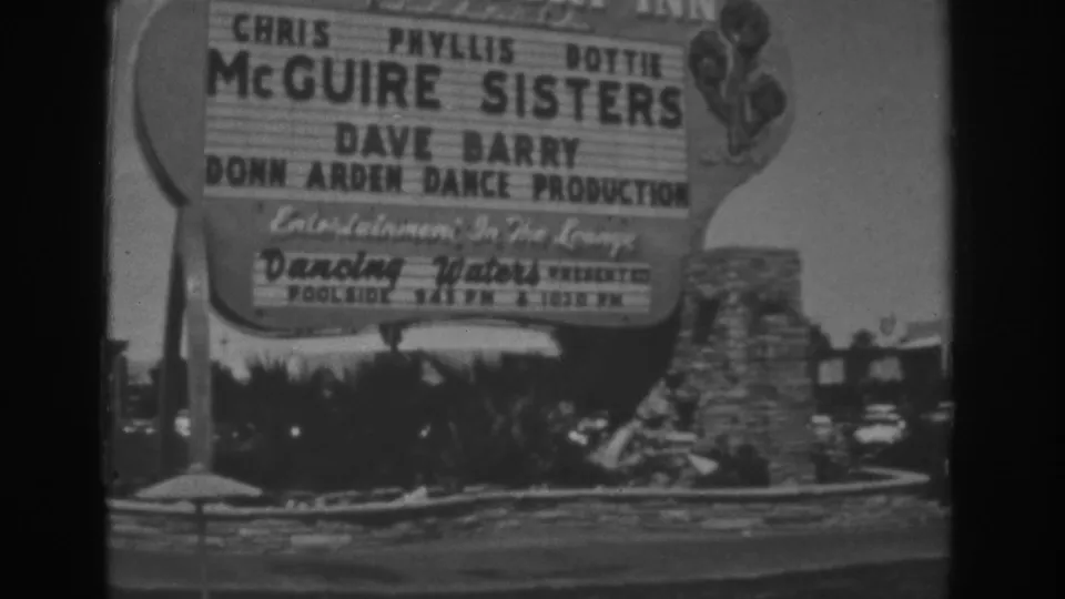 1962 Las Vegas frame focused on a roadside Stardust entertainment sign naming performers, with shrubs and stone landscaping below.
