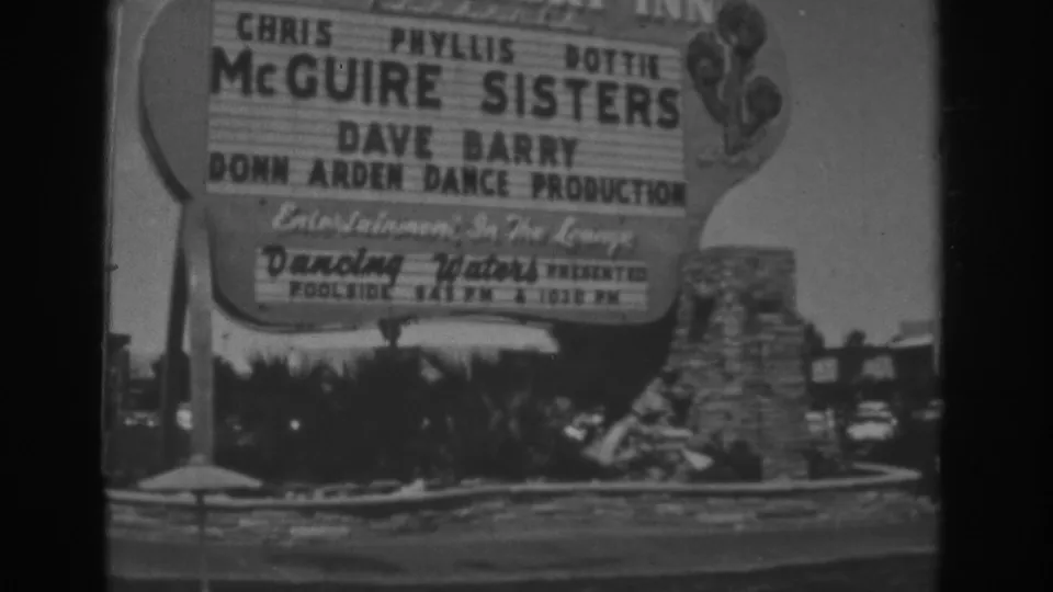 1962 Las Vegas frame holding on the Stardust roadside sign with performer listings, low landscaping, stone feature, and cars in the background.