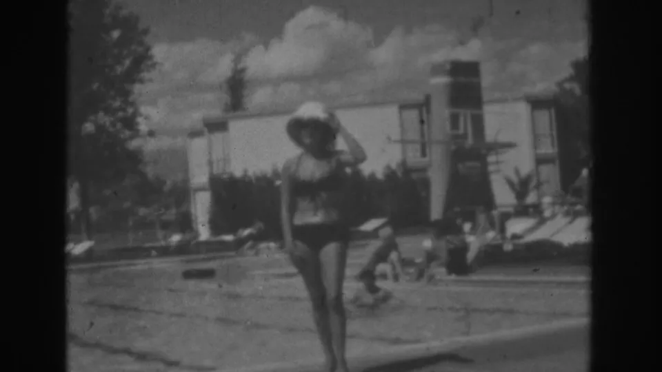 1962 Las Vegas poolside frame showing a woman in a swimsuit and broad hat walking past loungers and a building.
