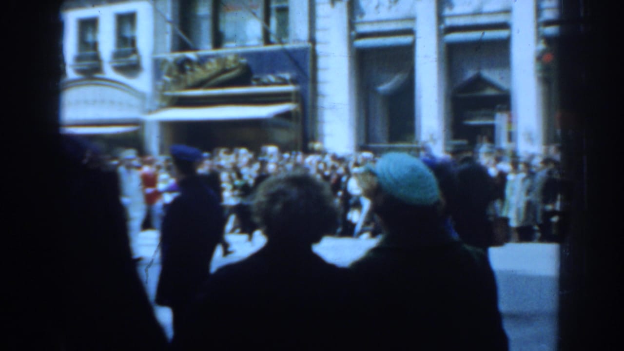 Sidewalk spectators watch a parade on a sunny city street with storefronts behind them.