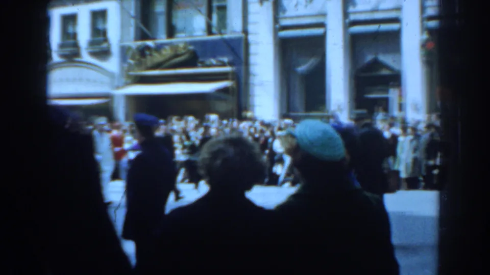 1962 New York City home-movie frame showing spectators in coats and hats watching a parade from the sidewalk.