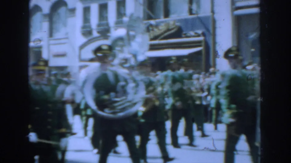 1962 New York City parade frame showing green-uniformed musicians marching past sidewalk spectators.
