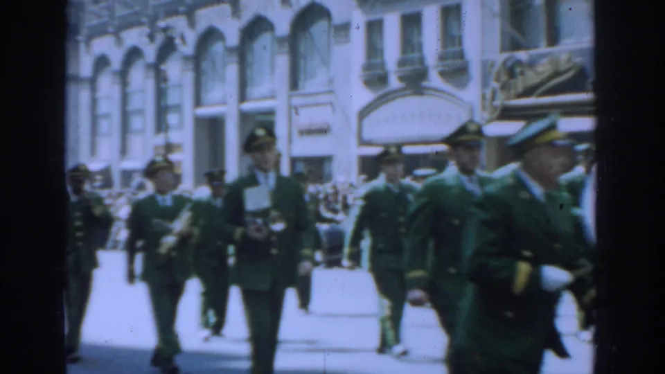 1962 New York City home-movie frame of green-uniformed parade band members moving past an urban storefront row.
