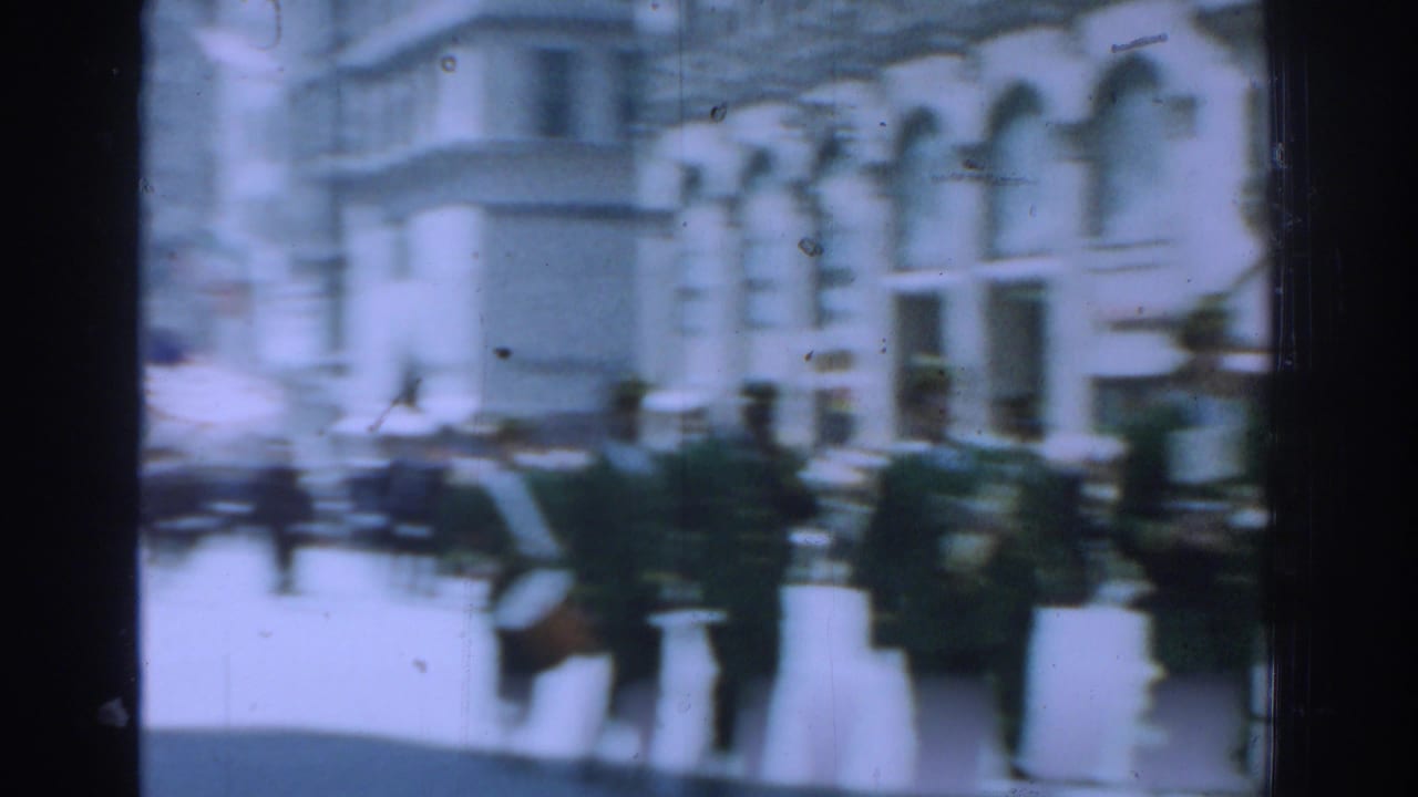 Marching personnel in green uniforms pass along a bright city street.