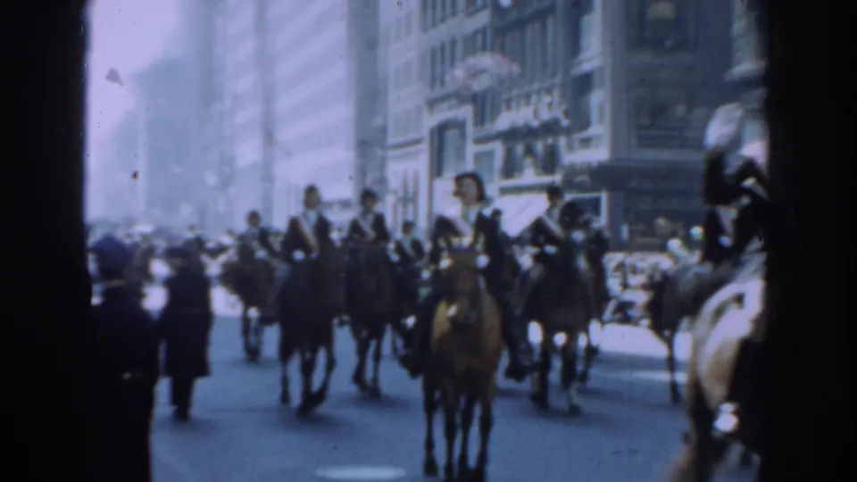 A group of riders on brown horses passes the camera in 1962 New York City parade footage, with spectators and tall buildings behind them.
