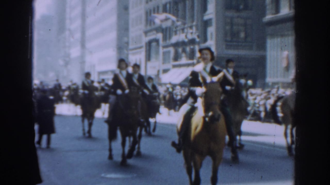 Mounted men in formal uniforms ride down a city street as spectators line the sidewalk.