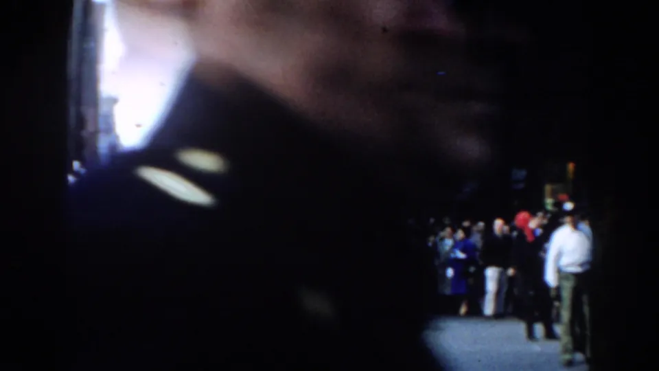 Obstructed 1962 New York City home-movie frame with a dark foreground figure and parade spectators visible in the street behind.