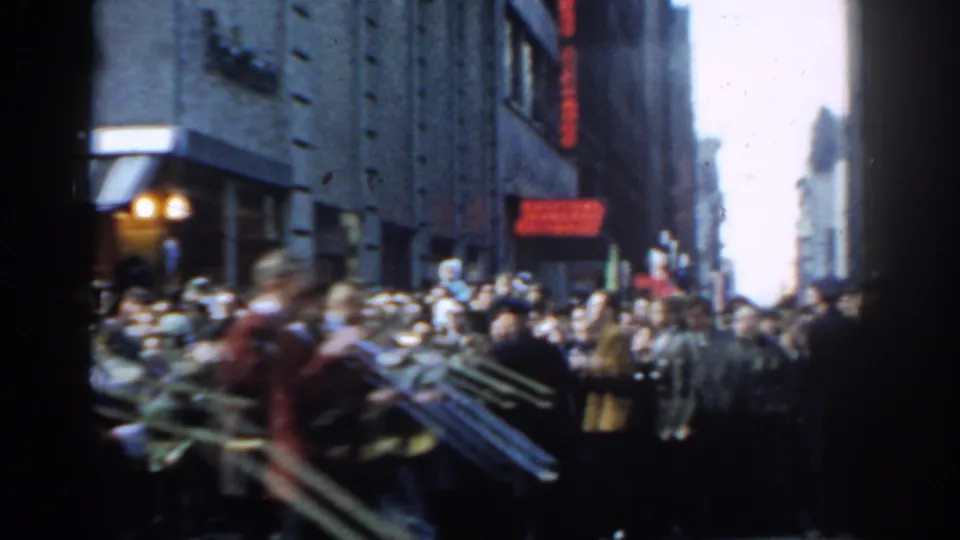1962 New York City parade frame showing blurred brass musicians moving past spectators along a tall-building corridor.