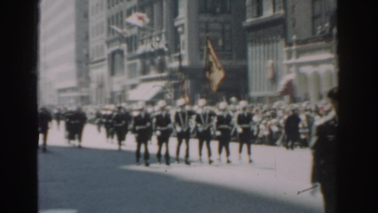 Uniformed marchers in white hats proceed along a city street before a sidewalk crowd.